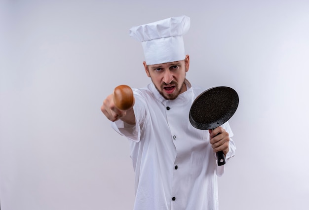 Chef maschio professionista cuoco in uniforme bianca e cappello da cuoco che tiene la padella che punta alla telecamera con la faccia arrabbiata in piedi sopra priorità bassa bianca