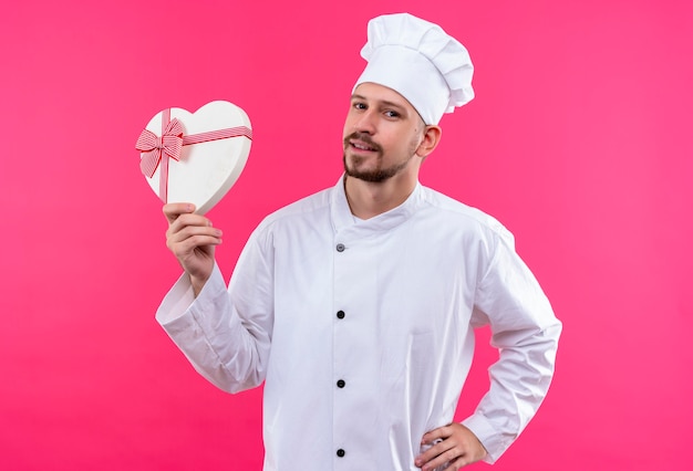 Профессиональный шеф-повар-мужчина в белой униформе и поварской шляпе держит подарочную коробку, уверенно улыбаясь на розовом фоне