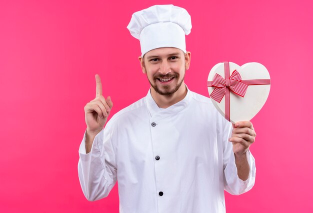Профессиональный шеф-повар-мужчина в белой униформе и поварской шляпе держит подарочную коробку, указывая пальцем вверх, весело улыбаясь на розовом фоне