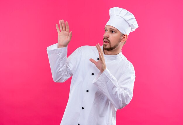 Professional male chef cook in white uniform and cook hat holding his hands up telling do not come closer over pink background