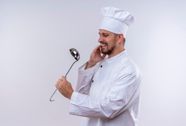 Professional male chef cook in white uniform and cook hat holding a ladle stressed and nervous biting nails standing over white background