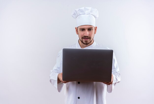 Professional male chef cook in white uniform and cook hat holding laptop looking at it intrigued standing over white background