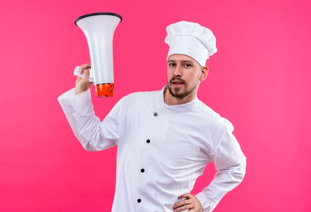 白い制服を着たプロの男性シェフが調理し、ピンクの背景の上に自信を持って立っているメガホンを保持している帽子を調理します。