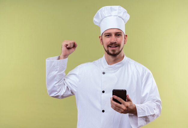 Professional male chef cook in white uniform and cook hat holding mobile phone raising fist happy and positive rejoicing his success standing over green background