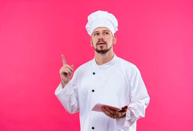 Professional male chef cook in white uniform and cook hat holding notebook looking up pointing finger up having new idea standing over pink background