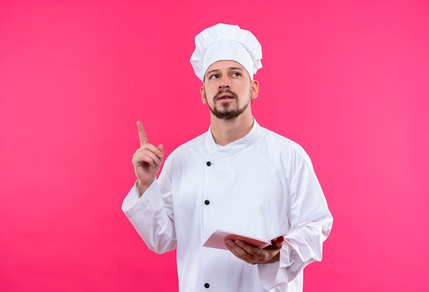 전문 남성 요리사 흰색 유니폼에 요리 하 고 분홍색 배경 위에 새로운 아이디어 서있는 데 가리키는 손가락을 찾고 노트북을 들고 요리 모자