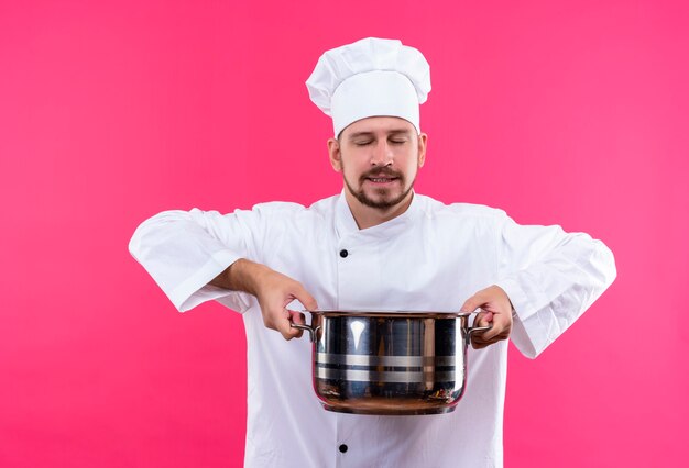 전문 남성 요리사는 흰색 유니폼을 입고 요리사 모자를 들고 분홍색 배경 위에 서있는 음식의 즐거운 냄새를 흡입합니다.