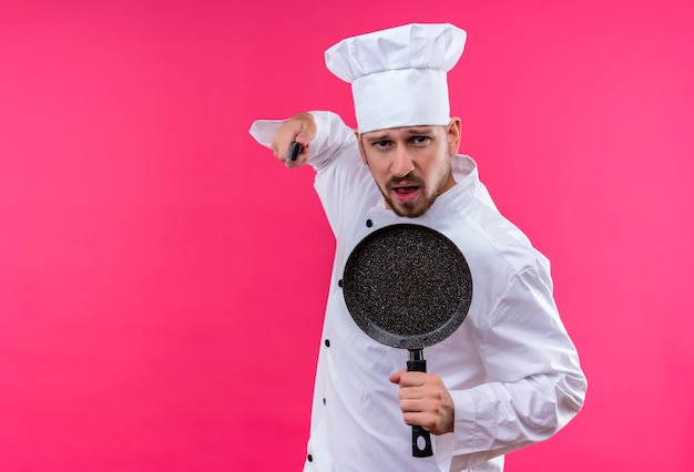 Professional male chef cook in white uniform and cook hat holding a pan and knife looking at camera threatening with a knife standing over pink background