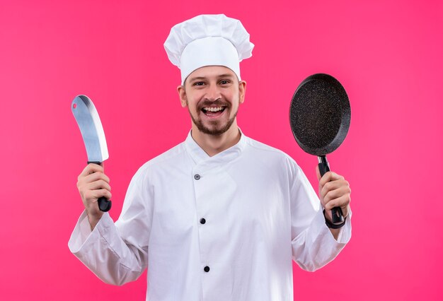 Professional male chef cook in white uniform and cook hat holding a pan and knife smiling cheerfully looking at camera standing over pink background