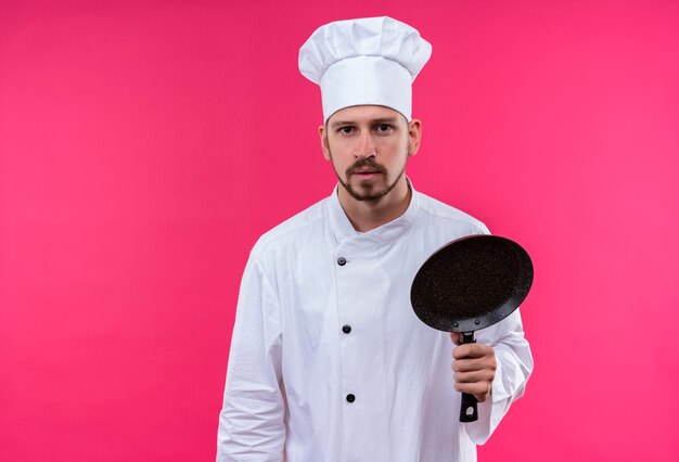 Chef maschio professionista cuoco in uniforme bianca e cappello da cuoco che tiene una padella che guarda l'obbiettivo con espressione seria in piedi su sfondo rosa