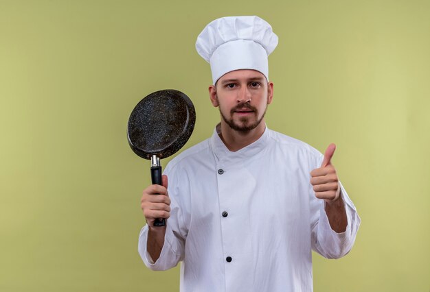 白い制服を着たプロの男性シェフが調理し、緑色の背景の上に立って親指を自信を持って見せて鍋を見ている帽子を調理します。