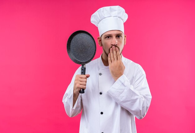 Professional male chef cook in white uniform and cook hat holding a pan looking surprised covering mouth with hand standing over pink background