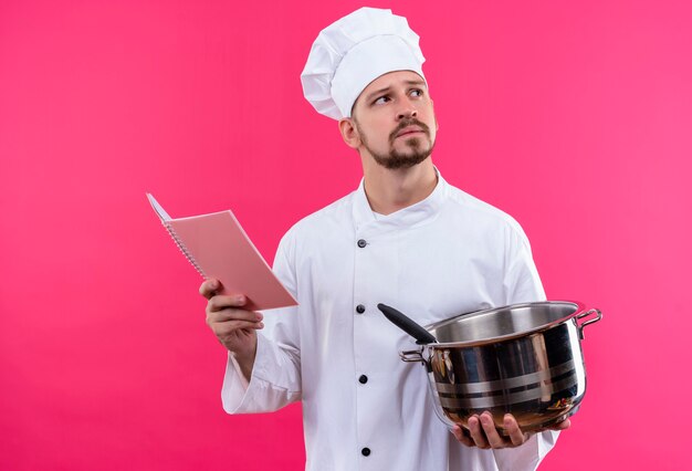 Профессиональный шеф-повар-мужчина в белой форме и поварской шляпе держит сковороду и блокнот, глядя вверх, думая, пытаясь сделать выбор, стоя на розовом фоне