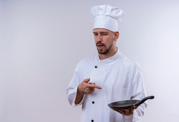 Professional male chef cook in white uniform and cook hat holding a pan pointing with finger to it looking at camera slyly standing over white background