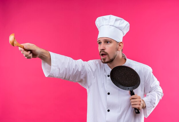 전문 남성 요리사 흰색 유니폼에 요리하고 분홍색 배경 위에 놀란 걱정 서 찾고 측면에 나무 숟가락으로 가리키는 냄비를 들고 요리 모자