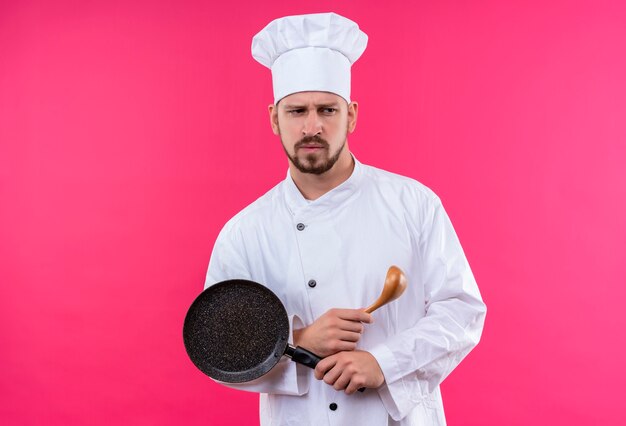 Профессиональный шеф-повар-мужчина в белой форме и поварской шляпе держит сковороду и деревянную ложку, недовольно смотрит в сторону и хмурится, стоя на розовом фоне