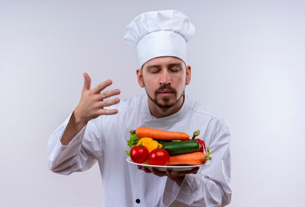 Professional male chef cook in white uniform and cook hat holding plate with vegetables inhales smell of them standing over white background