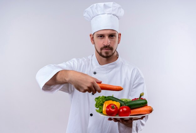 白い制服を着たプロの男性シェフが調理し、白い背景の上に立っている深刻な自信を持って式でカメラを見て野菜のプレートを保持している帽子を調理します。