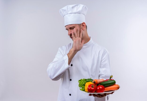 白い制服を着たプロの男性シェフが調理し、白い背景の上に立って手で防御ジェスチャーを作る野菜のプレートを保持している帽子を調理します。