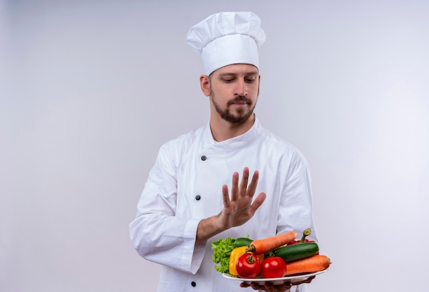 Professional male chef cook in white uniform and cook hat holding a plate with vegetables, making defense gesture with hand standing over white background