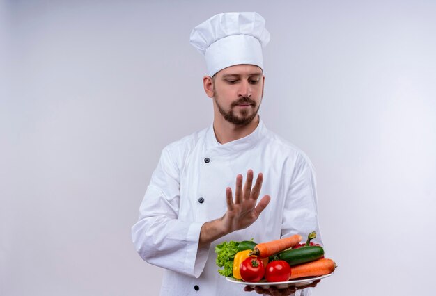 전문 남성 요리사는 흰색 유니폼을 입고 야채와 함께 접시를 들고 모자를 요리하고, 흰색 배경 위에 서있는 손으로 방어 제스처를 만듭니다.