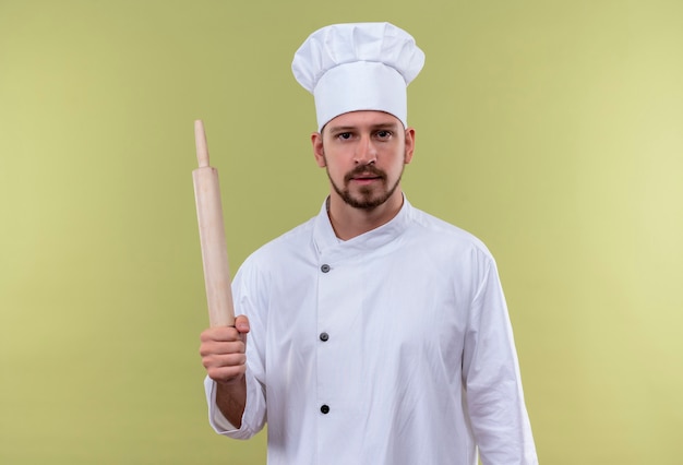 Professional male chef cook in white uniform and cook hat holding rolling pin looking confident standing over green background