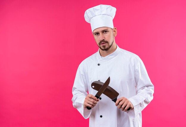 Professional male chef cook in white uniform and cook hat holding sharp knifes looking at camera with serious face standing over pink background