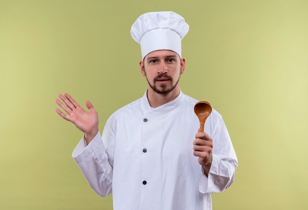 전문 남성 요리사 흰색 유니폼 요리와 녹색 배경 위에 자신감 서 찾고 손을 올리는 나무 숟가락을 들고 모자를 요리