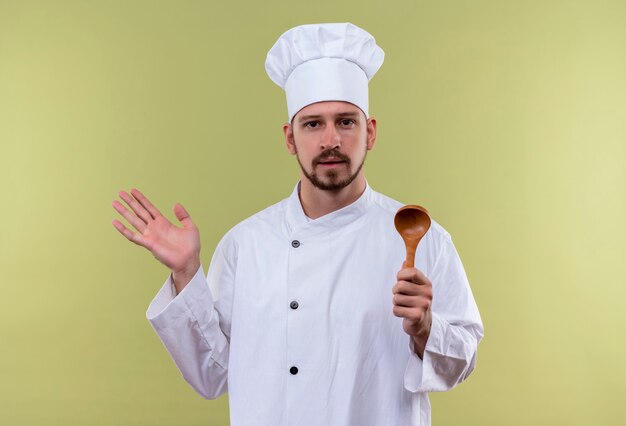 Профессиональный шеф-повар-мужчина в белой форме и поварской шляпе держит деревянную ложку, поднимая руку, уверенно глядя, стоя на зеленом фоне