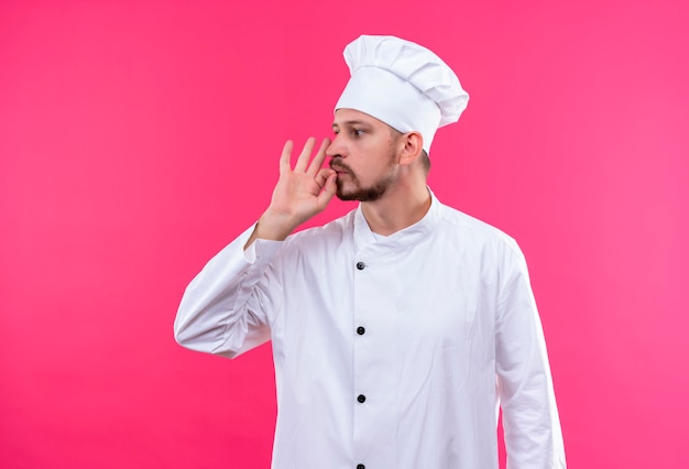 Professional male chef cook in white uniform and cook hat looking aside making silence gesture as closing his mouth with a zipper standing over pink background
