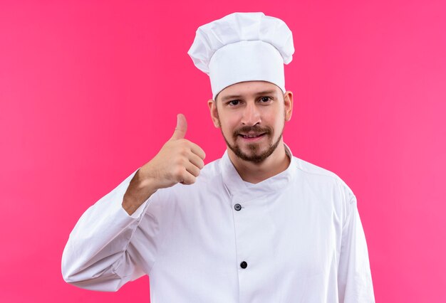 Professional male chef cook in white uniform and cook hat looking at camera smiling friendly showing thumbs up standing over pink background