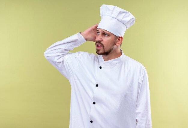 Professional male chef cook in white uniform and cook hat looking confused scratching his head for mistake standing over green background