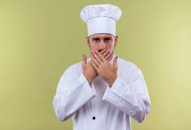 전문 남성 요리사 흰색 유니폼을 입고 요리 모자는 초 배경 위에 서있는 손으로 입을 덮고 놀란 찾고 요리