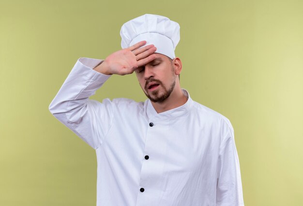 プロの男性シェフが白い制服を着て調理し、greeの背景の上に立って彼の頭に触れて疲れて過労探している帽子を調理します。