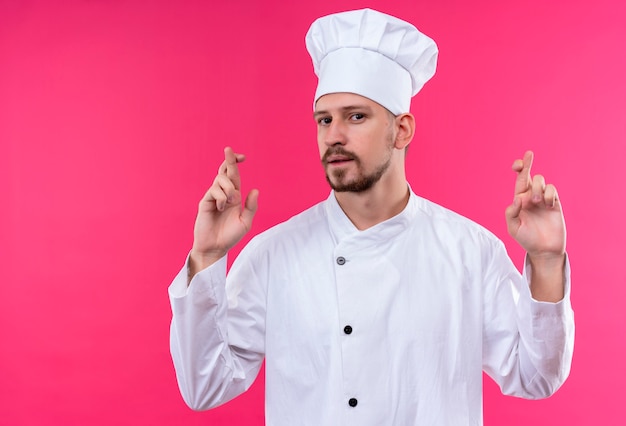 白い制服を着たプロの男性シェフが調理し、ピンクの背景の上に立って指を交差する望ましい願いを作る帽子を調理します。