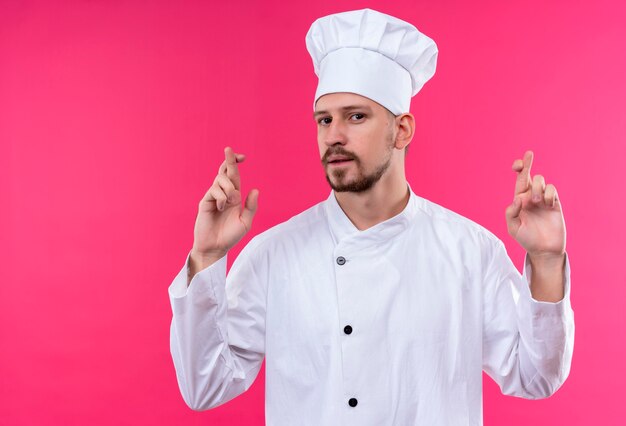 전문 남성 요리사는 흰색 유니폼을 입고 분홍색 배경 위에 서있는 바람직한 소원 횡단 손가락을 만드는 모자를 요리합니다.