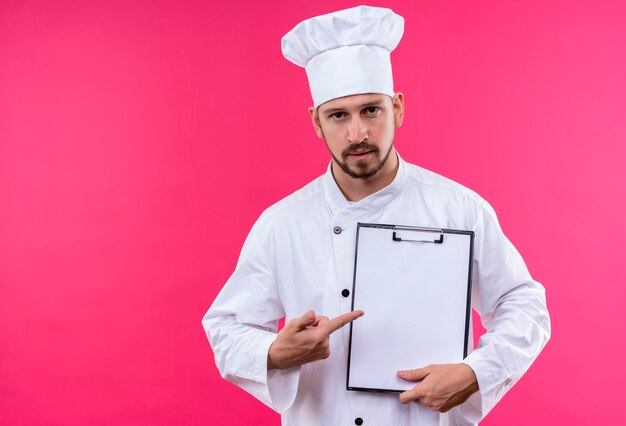 전문 남성 요리사는 흰색 유니폼을 입고 분홍색 배경 위에 서있는 빈 페이지 클립 보드를 제시하는 요리사 모자를 요리