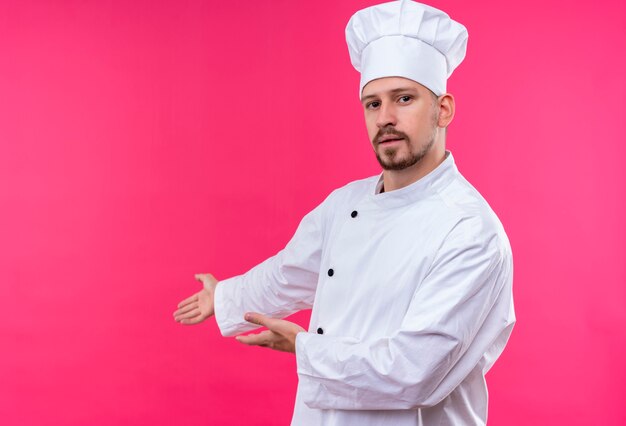 전문 남성 요리사는 흰색 유니폼을 입고 분홍색 배경 위에 서있는 그의 손의 팔로 복사 공간을 제시하는 모자를 요리합니다.