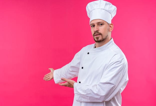 Professional male chef cook in white uniform and cook hat presenting copy space with arms of his hands standing over pink background