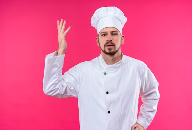 Professional male chef cook in white uniform and cook hat raising hand as asking question standing over pink background