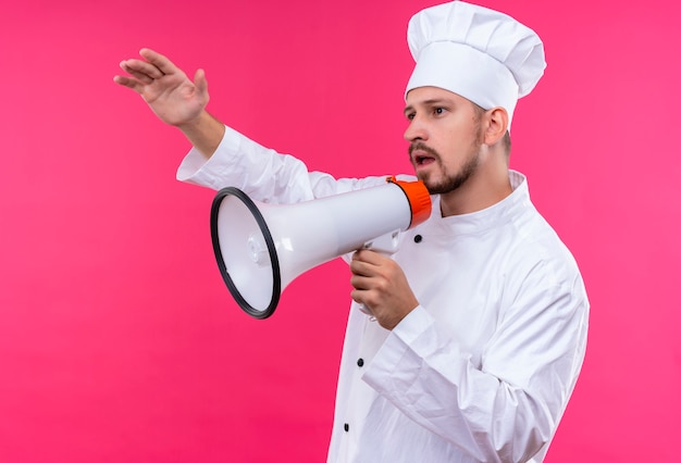 Professional male chef cook in white uniform and cook hat speaking to megaphone calling someone waving with hand standing over pink background