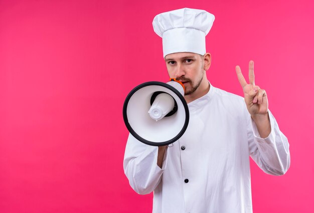 白い制服を着たプロの男性シェフが調理し、ピンクの背景の上に立っている数2を示すメガホンに話す帽子を調理します。