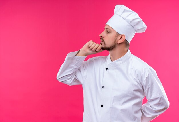전문 남성 요리사는 흰색 유니폼을 입고 분홍색 배경 위에 생각하는 턱에 손으로 옆으로 서있는 모자를 요리합니다.