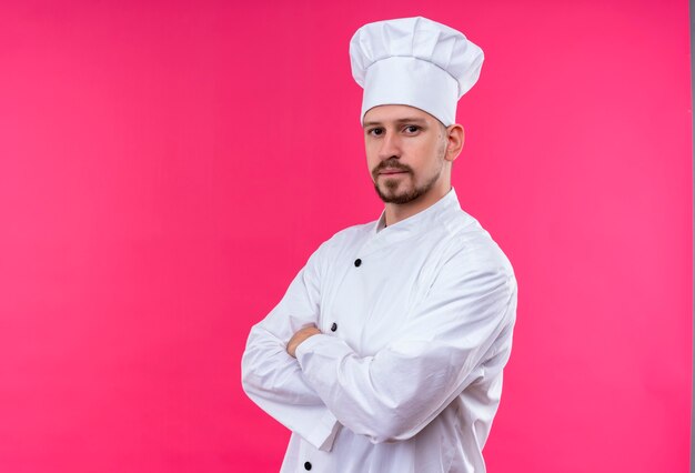 Professional male chef cook in white uniform and cook hat standing with arms crossed looking confident over pink background