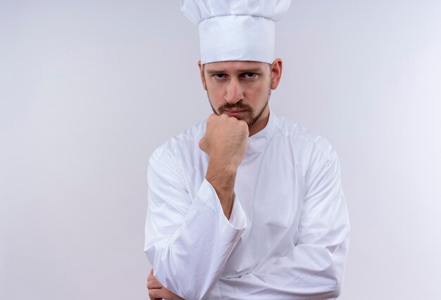 Профессиональный шеф-повар-мужчина в белой форме и поварской шляпе, стоя с кулаком на подбородке с задумчивым выражением лица на белом фоне