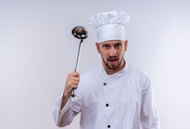 Professional male chef cook in white uniform and cook hat swinging a ladle with angry face standing over white background