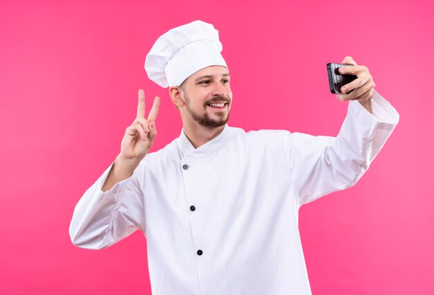 Professional male chef cook in white uniform and cook hat taking selfie using his smartphone showing victory sign smiling standing over pink background