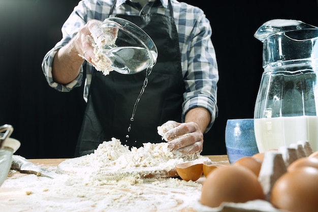 Professional male cook sprinkles dough with flour, preapares or bakes bread or pasta at kitchen table, has dirty uniform, isolated over black chalk background. Baking concept