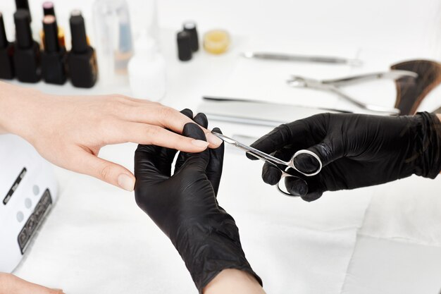 Professional manicurist in black gloves cutting cuticle with manicure scissors.