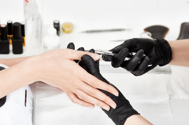 黒い手袋をしたプロのネイリストが、マニキュアキューティクルトリマーでマニキュアを作るクライアントにサービスを提供します。